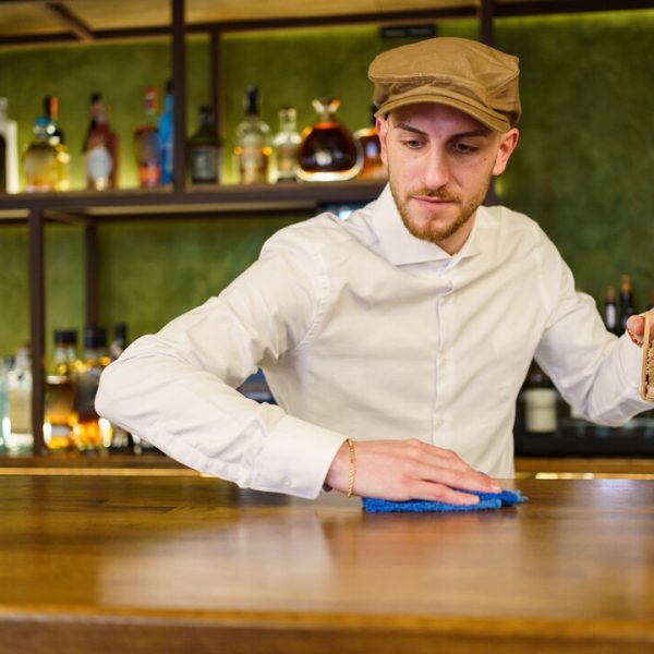 Bearded man in white shirt and cap removing napkin holder and wiping counter with cloth while working in bar in evening