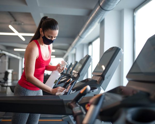 Fitness woman instructor with face mask disinfecting equipment in gym, coronavirus concept.