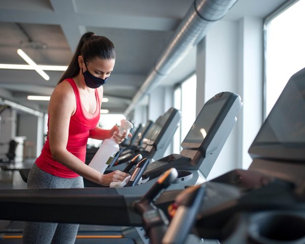 Fitness woman instructor with face mask disinfecting equipment in gym, coronavirus concept.