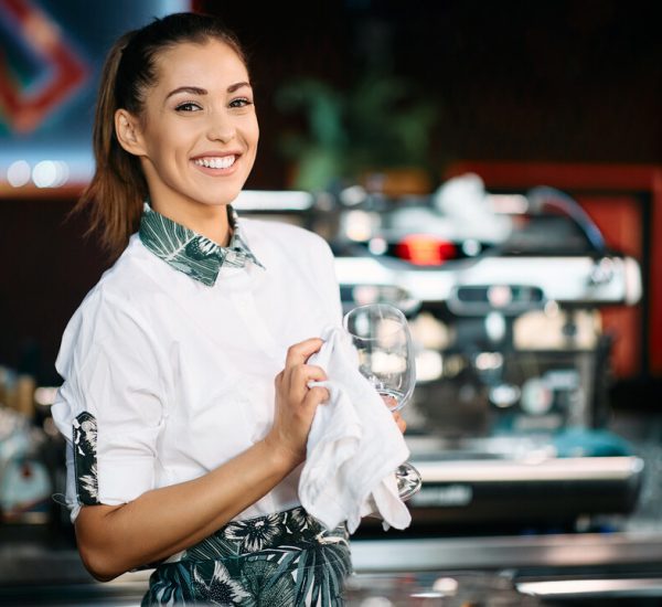 Young happy woman cleaning drinking glasses while working as barista in a pub.