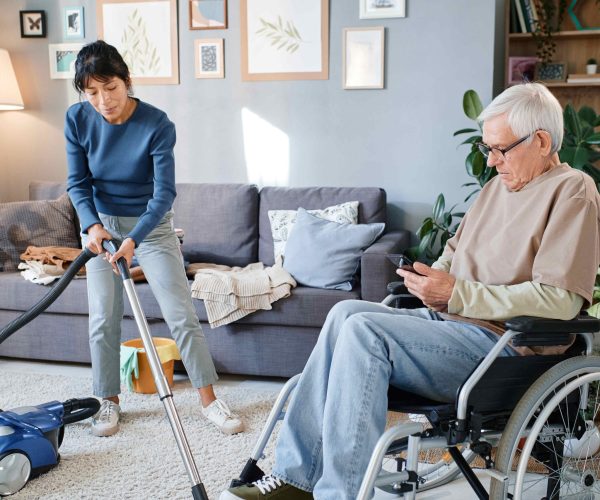 Senior man using mobile phone while housekeeper helping him to clean the room with vacuum cleaner