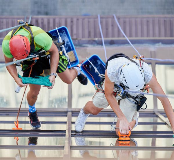 Industrial mountaineering. Top view of professional cleaners hanging on ropes while cleaning skyscraper windows. Two men window washers using cleaning tools while washing building facade.